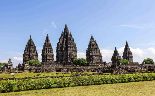 Die Tempelanlage Prambanan in Yogyakarta, Indonesien, besteht aus mehreren hohen, dunklen Steintürmen mit detailreichen Verzierungen. Vor der Anlage erstreckt sich eine gepflegte Grünfläche. Der Himmel ist blau mit wenigen weißen Wolken.
