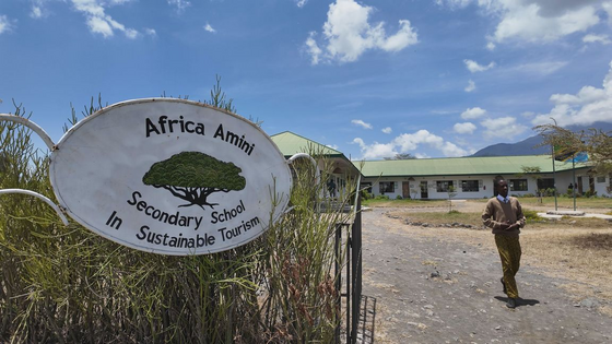 Das Africa-Amini-Projekt Links im Bild sieht man ein Schild für die Africa Amini Secondary School, rechts im Hintergrund die Schulgebäude und im Mittelgrund einen Afrikaner, der über einen Weg läuft.