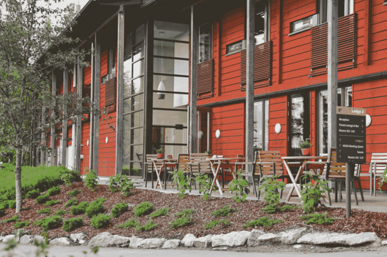 Man sieht die Fassade eines zweistöckigen Gebäudes, die in rotem Holz gestaltet ist. Auf der Terrasse vor dem Haus rechts im Bild stehen einige Holztische und -stühle.
