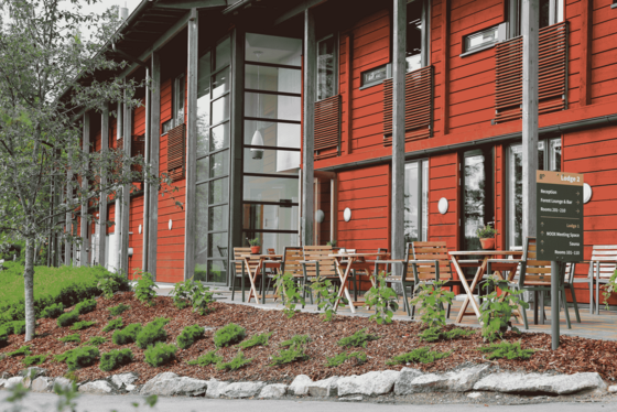 Man sieht die Fassade eines zweistöckigen Gebäudes, die in rotem Holz gestaltet ist. Auf der Terrasse vor dem Haus rechts im Bild stehen einige Holztische und -stühle.