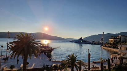 Entlang des vorderen Bildrands zieht sich eine palmengesäumte Promenade. In der mit Hügeln umfassten Bucht liegen mehrere Boote. Über den Hügeln im Hintergrund steht die Sonne sehr tief, der Himmel ist blau und wolkenlos. Die untergehende Sonne taucht die Promenade in rosa Abendlicht.