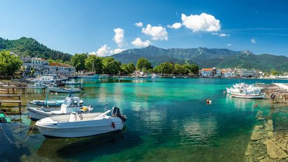 Thassos: Hafen der Inselhauptstadt Limenas Man blickt über eine Bucht mit klarem türkisfarbenen Wasser, in der Bötchen dümpeln. Auf der gegenüberliegenden Seite der Bucht befinden sich Gebäude mit dazwischenstehenden Bäumen, dahinter erheben sich ebenfalls baumbestandene Hügel und darüber ein strahlend blauer Himmel.