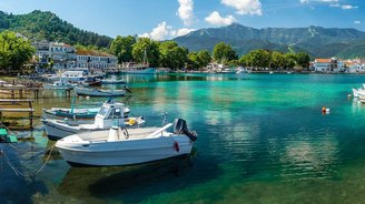 Man blickt über eine Bucht mit klarem türkisfarbenen Wasser, in der Bötchen dümpeln. Auf der gegenüberliegenden Seite der Bucht befinden sich Gebäude mit dazwischenstehenden Bäumen, dahinter erheben sich ebenfalls baumbestandene Hügel und darüber ein strahlend blauer Himmel.