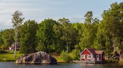 An einem Seeufer steht ein rotes Holzhaus umgeben von Bäumen.
