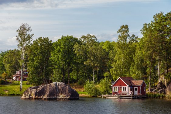 An einem Seeufer steht ein rotes Holzhaus umgeben von Bäumen.