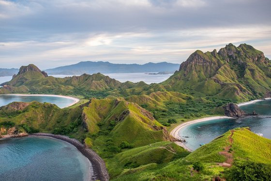 Eine hügelige Landschaft mit grasbedeckten Bergen erstreckt sich über mehrere Inseln im Komodo-Nationalpark. Die Küsten sind von geschwungenen Sandstränden umgeben. Im Hintergrund ist das offene Meer mit weiteren Inseln sichtbar.