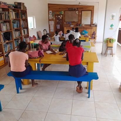 Man schaut in einen Raum, in dem hintereinander drei große, gelbe Tische stehen. An den Tischen sitzen junge Afrikanerinnen und lesen. An der linken Wand steht ein großes Bücherregal.