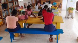 Man schaut in einen Raum, in dem hintereinander drei große, gelbe Tische stehen. An den Tischen sitzen junge Afrikanerinnen und lesen. An der linken Wand steht ein großes Bücherregal.