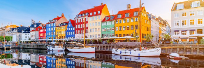 Bunte Häuserreihe, die sich im Fluss spiegeln. Davor befinden sich angelegte Boote.