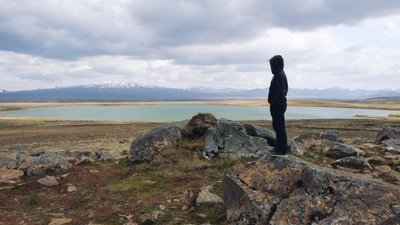 Person blickt über See- und Berglandschaft am Uxahryggir Eine in Schwarz gekleidete Person steht auf einem Felsen und schaut über eine weite Ebene mit See. Am Horizont liegt ein schneebedeckter Gebirgszug unter dramatischem Himmel.