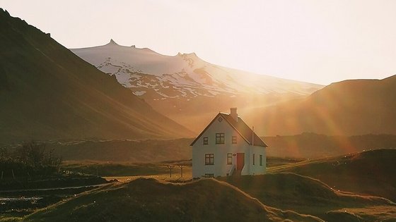 Haus in Arnarstapi im Licht der Mitternachtssonne Ein einsam stehendes, weißes Haus liegt eingebettet in einer moosigen Hügellandschaft. Im Hintergrund erhebt sich ein schneebedeckter Berg, beleuchtet vom goldenen Licht der untergehenden Mitternachtssonne.