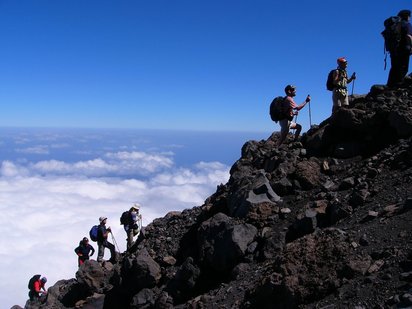 Aufstieg zum Pico do Fogo Diagonal von links unten nach rechts oben zieht sich ein Bergkamm mit einer Gruppe von Wanderern. Die Gruppe läuft über einem Meer aus weißen Wolken, dahinter der blaue Himmel.