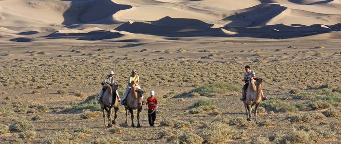 Durch eine Steppe mit Sanddünen reiten Touristen auf Kamelen