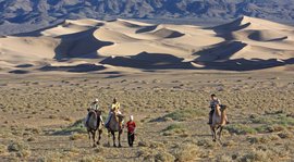 Durch eine Steppe mit Sanddünen reiten Touristen auf Kamelen