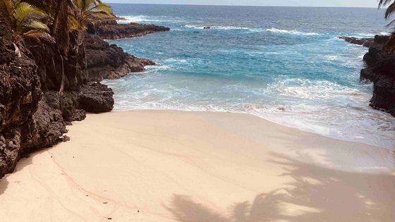 Ansicht einer Bucht, in der links und rechts die dunklen Felswände den weißen Sandstrand umranden. Im Hintergrund ist das blaue Meer.