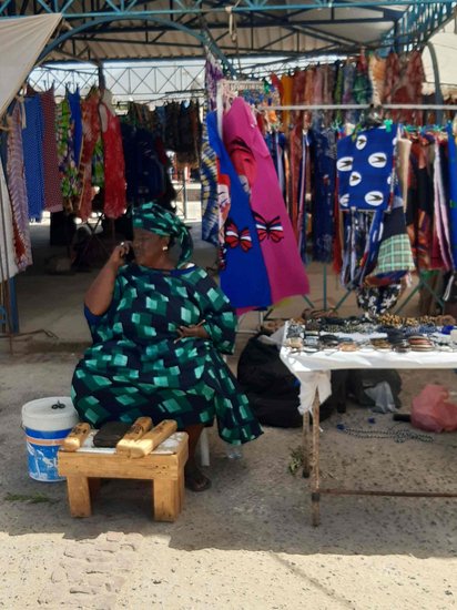 Eine Afrikanerin sitzt vor einem Marktstand mit bunter Kleidung.