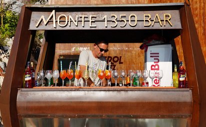 Die gesamte Bildfläche wird von einem hölzernen Bar-Stand ausgefüllt, hinter dem ein Barkeeper Longdrinks zubereitet. Vor ihm auf der Theke stehen einige mit Aperol Spritz und anderen Getränken gefüllte Kelche, einige sind noch leer.