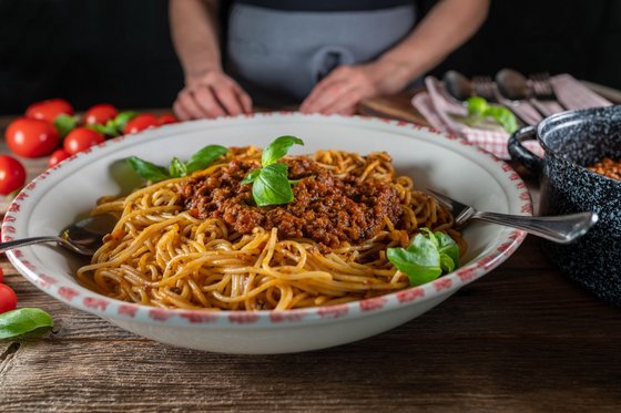 Im Vordergrund steht eine Schüssel Spaghetti mit Fleischsoße und Basilikumblättern, im Hintergrund eine Person und frische Tomaten auf dem Tisch.