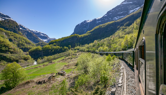 Mit der Dovrebahn durch Norwegens Norden Aufnahme aus einem Zugfenster entlang des Zuges, der durch eine gebirgige im unteren Teil grüne, im oberen Teil felsig-schneeige Landschaft fährt.