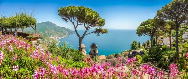Über ein Blütenmeer im Vordergrund schaut man den Hang hinab auf den Golf von Salerno an der Amalfiküste.