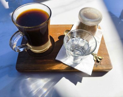 Auf einem Holzbrettchen steht ein Kaffee in einem Glas mit Henkel, daneben ein Wasserglas, brauner Zucker in einem Deckelglas und ein kleiner goldener Löffel.