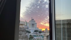 Man schaut aus einem geöffneten Fenster hinaus auf das Nationaldenkmal Viktor Emanuels II. Der fahlblaue Himmel beginnt in der Abenddämmerung orange zu leuchten.