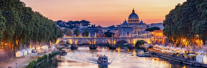 Das Bild zeigt eine malerische Aussicht auf Rom bei Sonnenuntergang. Im Vordergrund sind historische Gebäude zu sehen, darunter der imposante Petersdom.
