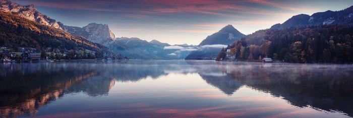 Panoramaaufnahme eines Bergsees in den österreichischen Alpen, umgeben von grünen Wiesen und imposanten Gipfeln, die sich im klaren Wasser spiegeln.
