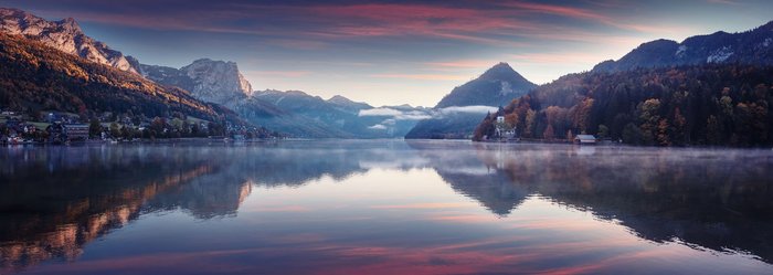 Panoramaaufnahme eines Bergsees in den österreichischen Alpen, umgeben von grünen Wiesen und imposanten Gipfeln, die sich im klaren Wasser spiegeln.