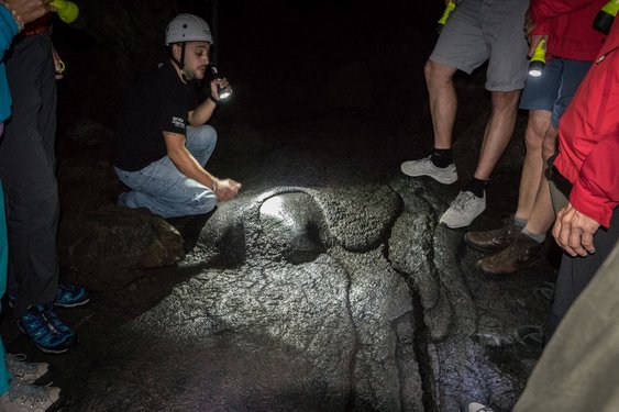 Man sieht den Boden einer Höhle aus erkalteter Lava. Ein von mehreren Menschen umstandener Führer leuchtet mit einer Taschenlampe auf eine Stelle im Boden und scheint etwas zu erklären.