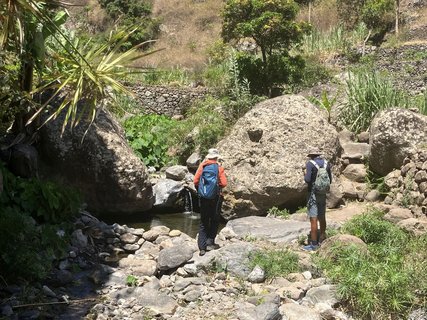 Zwei Männer mit Wanderrucksäcken und Kopfbedeckung stehen vor einem von Geröll und grüner Vegetation umgebenen Wasserloch.