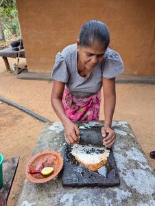 Eine Sri-Lankerin steht hinter einem Tisch und verarbeitet auf einem Brett mit einem Mörser verschiedene Zutaten zu einer Paste. Links auf dem Tisch befindet sich eine Schale aus Ton mit Chilis und einer Zitronenhälfte. Der Hintergrund wird von einer braunen Hauswand gebildet, an de3r man links vorbei in einen grünen Garten schaut.