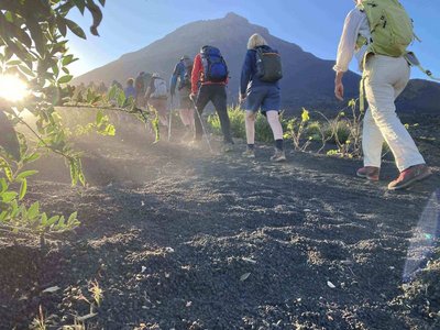 Eine Gruppe Wanderer bewegt sich im Gänsemarsch über Lavaasche von rechts nach links bergan auf dem Pico do Fogo. Mittig im Bildhintergrund sieht man den Gipfel des Vulkans.