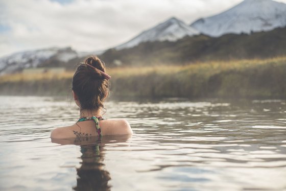 Auf dem Bild ist eine Frau zu sehen, die in einer heißen Quelle mitten in der Natur von Island badet.