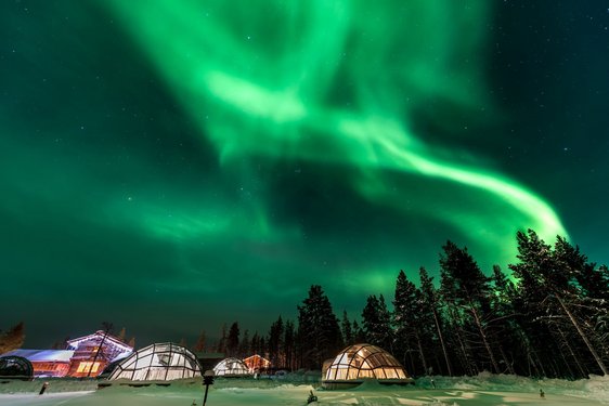 Am unteren Bildrand steht ein erleuchtetes Haus und mehrere große Igluzelte; darüber leuchtet der Himmel unter einem grünen Polarlicht.