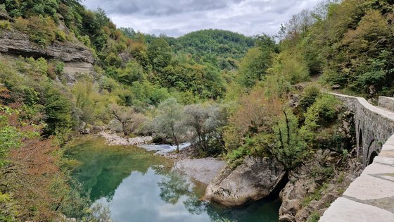 Das Foto wurde von einer Brücke über ein Flüsschen aus aufgenommen, die sich rechts im Bild weiter fortsetzt. Der Flusslauf wird von grün bewachsenen Hügeln flankiert. Oben ist ein schmaler Streifen grauer Himmel zu sehen.