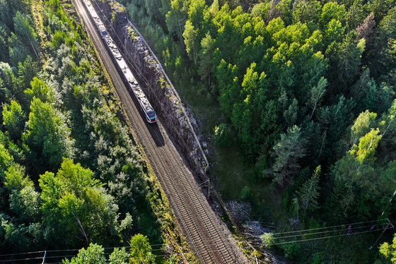 Mit dem Zug durch Finnlands Natur Man blickt aus der Aufsicht auf einen Wald, der von links oben nach vorne mittig von Schienen geteilt wird. von links oben fährt ein Zug in das Bild.
