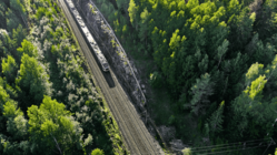 Man blickt aus der Aufsicht auf einen Wald, der von links oben nach vorne mittig von Schienen geteilt wird. von links oben fährt ein Zug in das Bild.