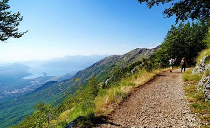 Die rechte vordere Bildhälfte wir von einem Wanderweg eingenommen, der rechts im Bildmittelgrund um eine Kurve führt. Dort, wo er aus dem Blick entschwindet, laufen zwei Wanderer vom Betrachter weg. Links des Wegs fällt der grüne Hang steil bergab hin zu einem See links im Bildmittelgrund.