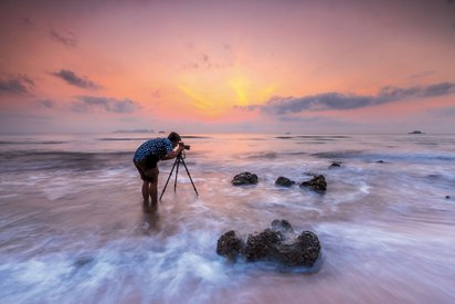 Fotografieren am Meer Ein Mann steht bis zu den Waden im Meer und schaut durch eine Kamera, die auf einem Stativ befestigt ist.