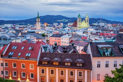 Die Altstadt von Linz Über die Dächer der Altstadt von Linz, aus der sich zwei Kirchen erheben, schaut man auf die Stadt im Dämmerlicht. Dahinter zieht sich eine Hügellandschaft entlang des Horizonts.