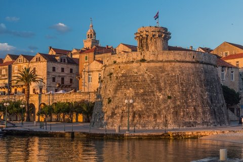 Ein alter Steinturm in Korcula, Kroatien, mit Blick auf das blaue Meer. Im Hintergrund leuchten die roten Ziegeldächer der Altstadt in der Sonne.