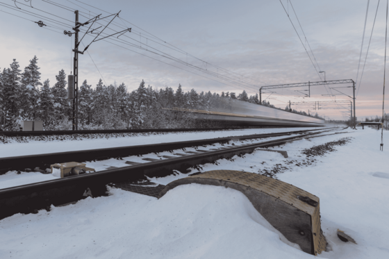 Die Bildmitte wird von Gleisen durchzogen, auf denen eine Zug mit hoher Geschwindigkeit vorüber rauscht. Hinter den Schienen stehen schneebedeckte Bäume, der Bahndamm davor ist ebenfalls schneebedeckt.